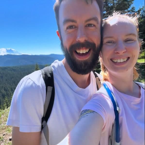 Julia and RJ taking a selfie on a sunny hike, with mountains and an evergreen ridge behind them.